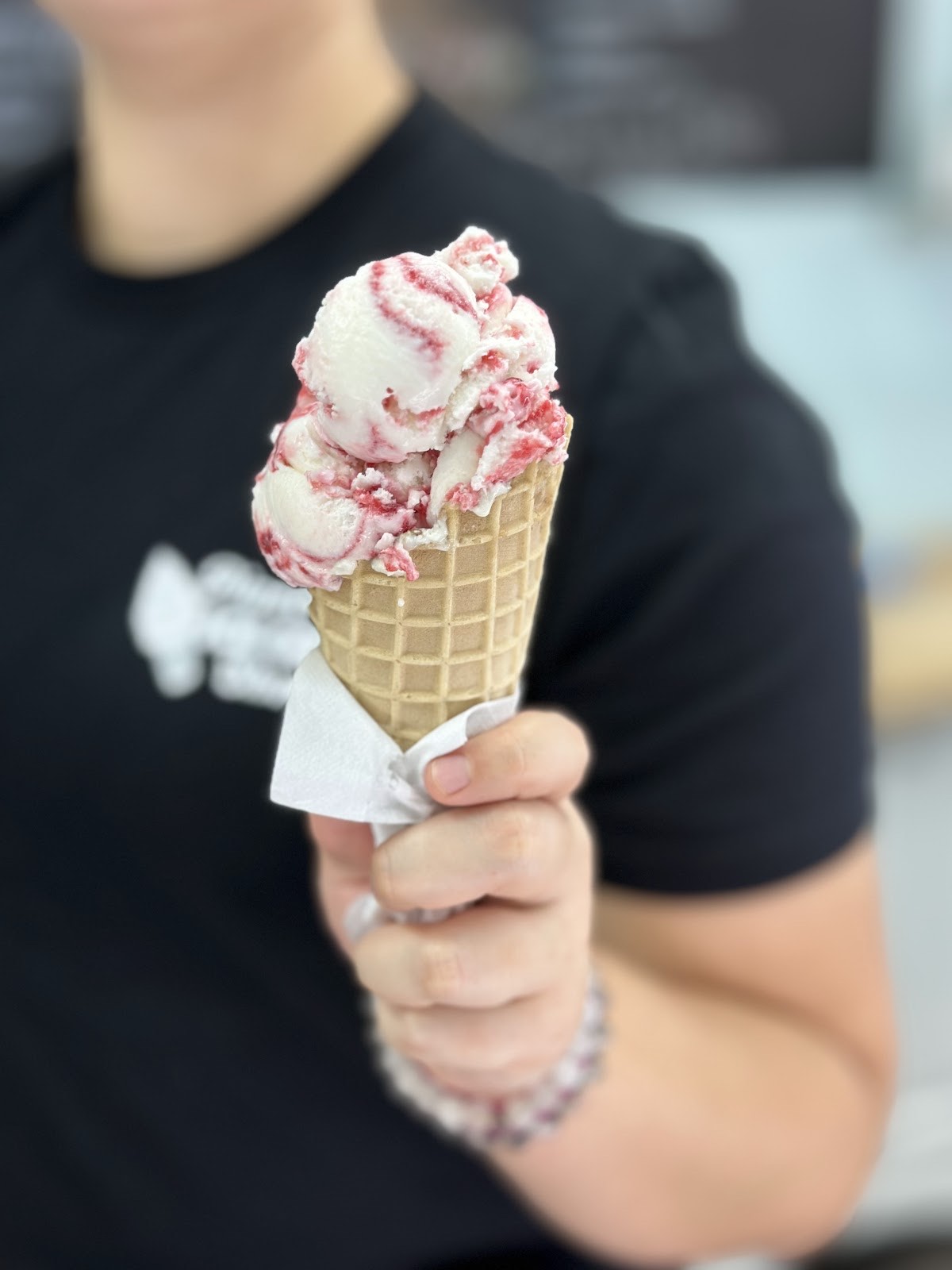 A hand holding a waffle cone with two scoops of creamy strawberry-swirl homemade ice cream at Pinecraft Ice Cream Shoppe